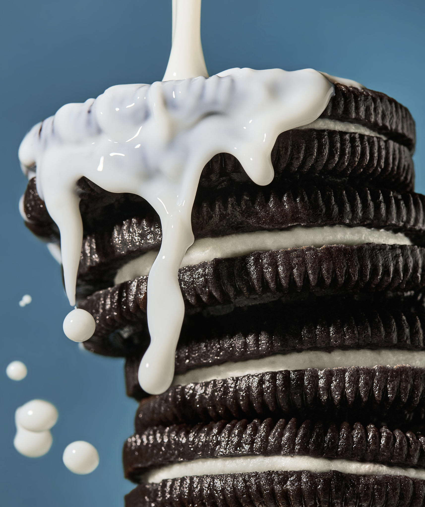 Close-up of a stacked chocolate sandwich cookies with white cream filling, drizzled with milk against a solid blue background.