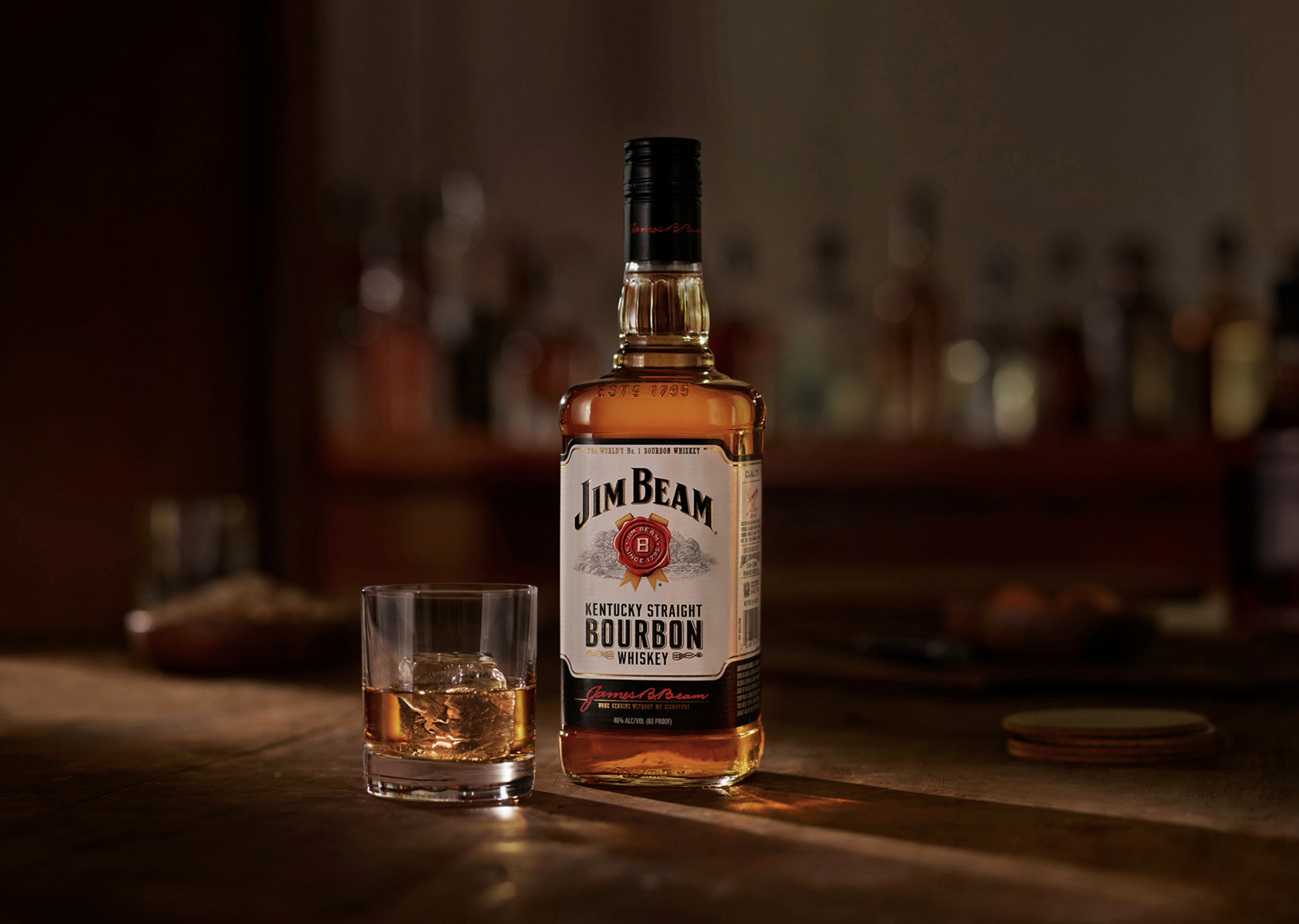 Jim Beam bourbon whiskey bottle and glass of whiskey with ice on a wooden bar surface in a warm, inviting setting.