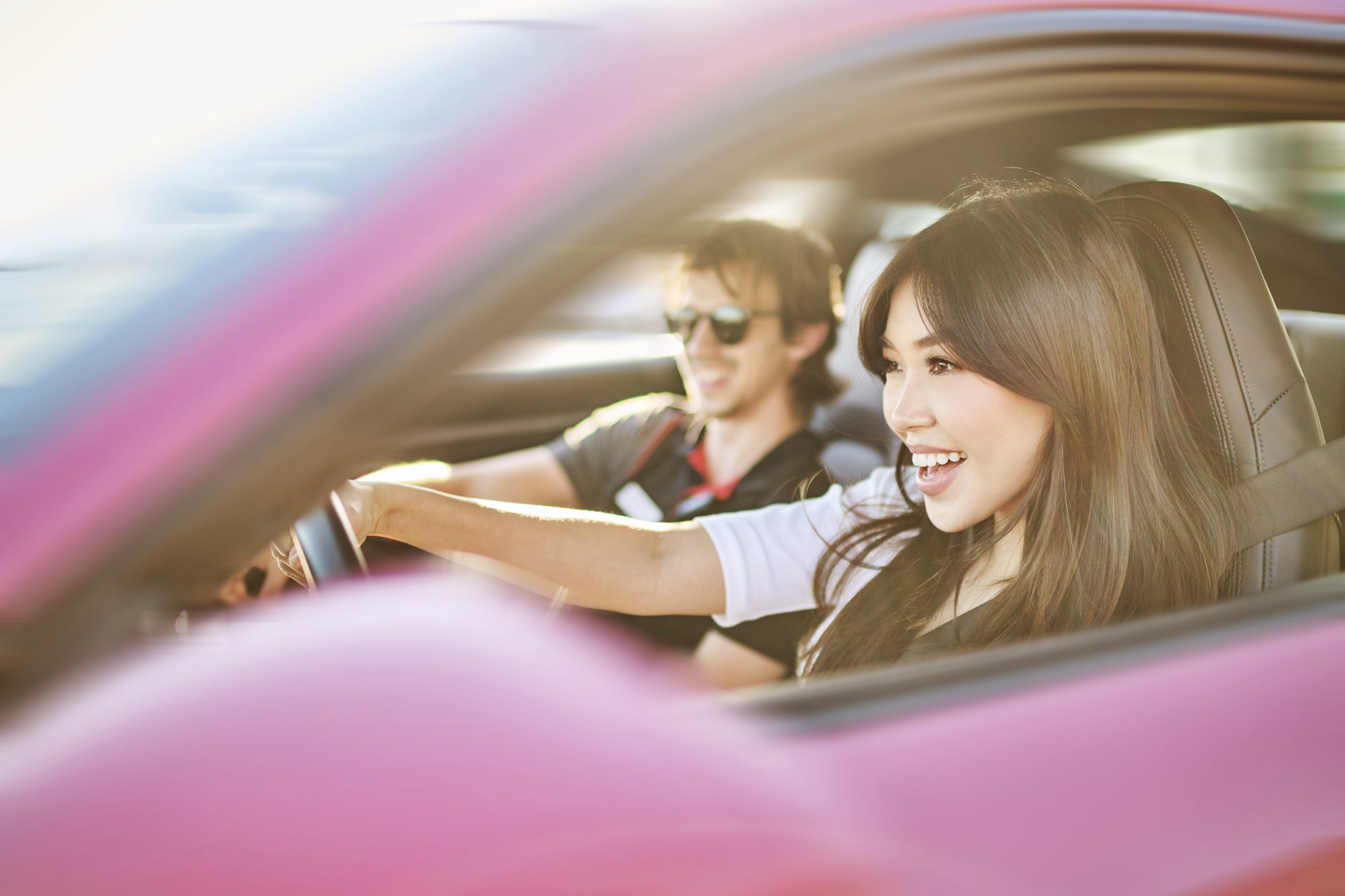 Young woman and man smiling and driving in a pink convertible car on a sunny day. Interior shot of two happy people.