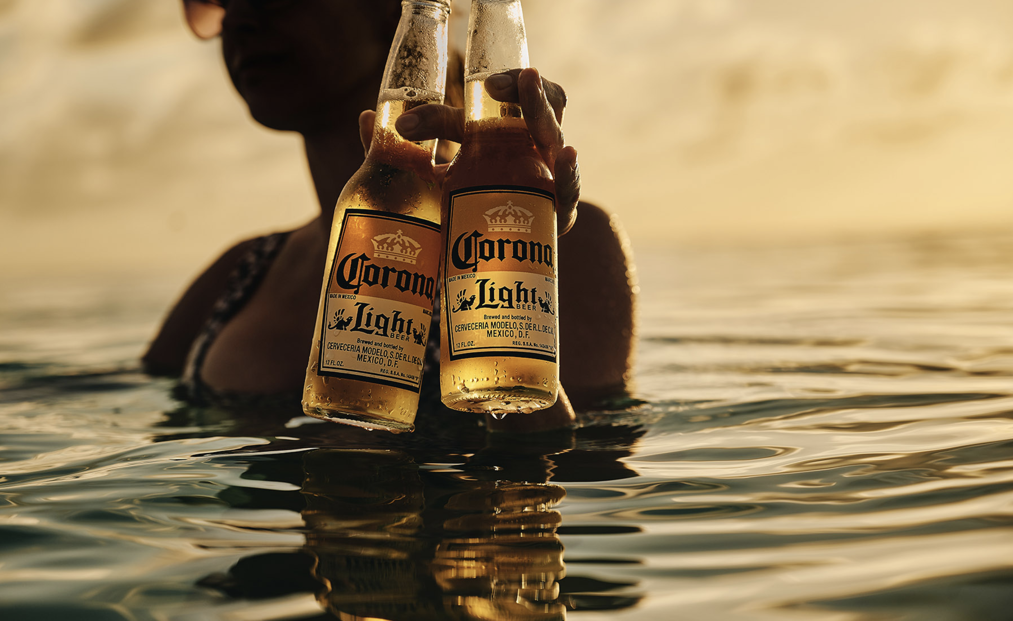 Two Corona Light beer bottles are held by a woman in the ocean at sunset with golden light reflecting on the water and glass bottles.
