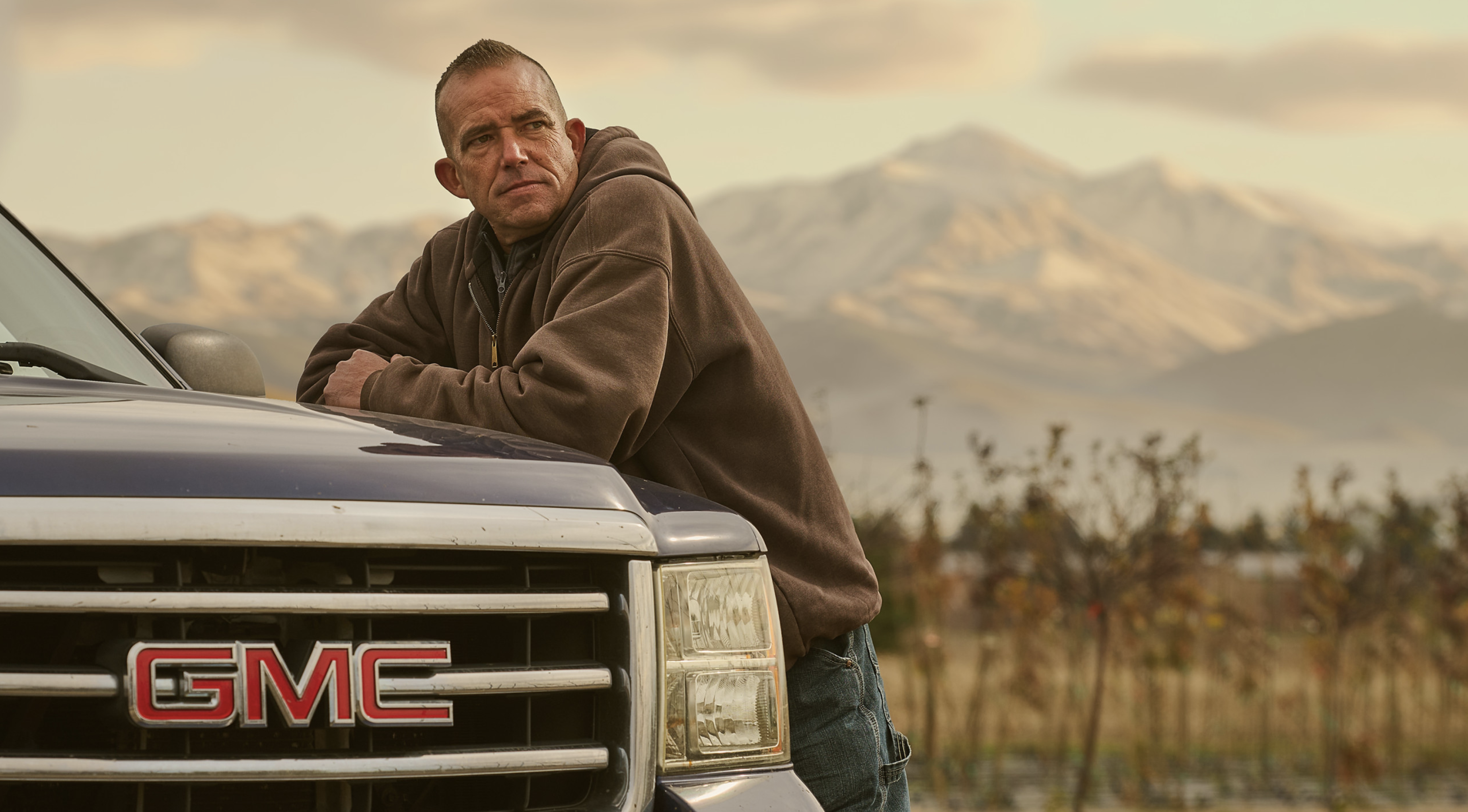 Adult male leans against a blue GMC pickup truck with a mountain backdrop. The man is wearing a brown hoodie and jeans.