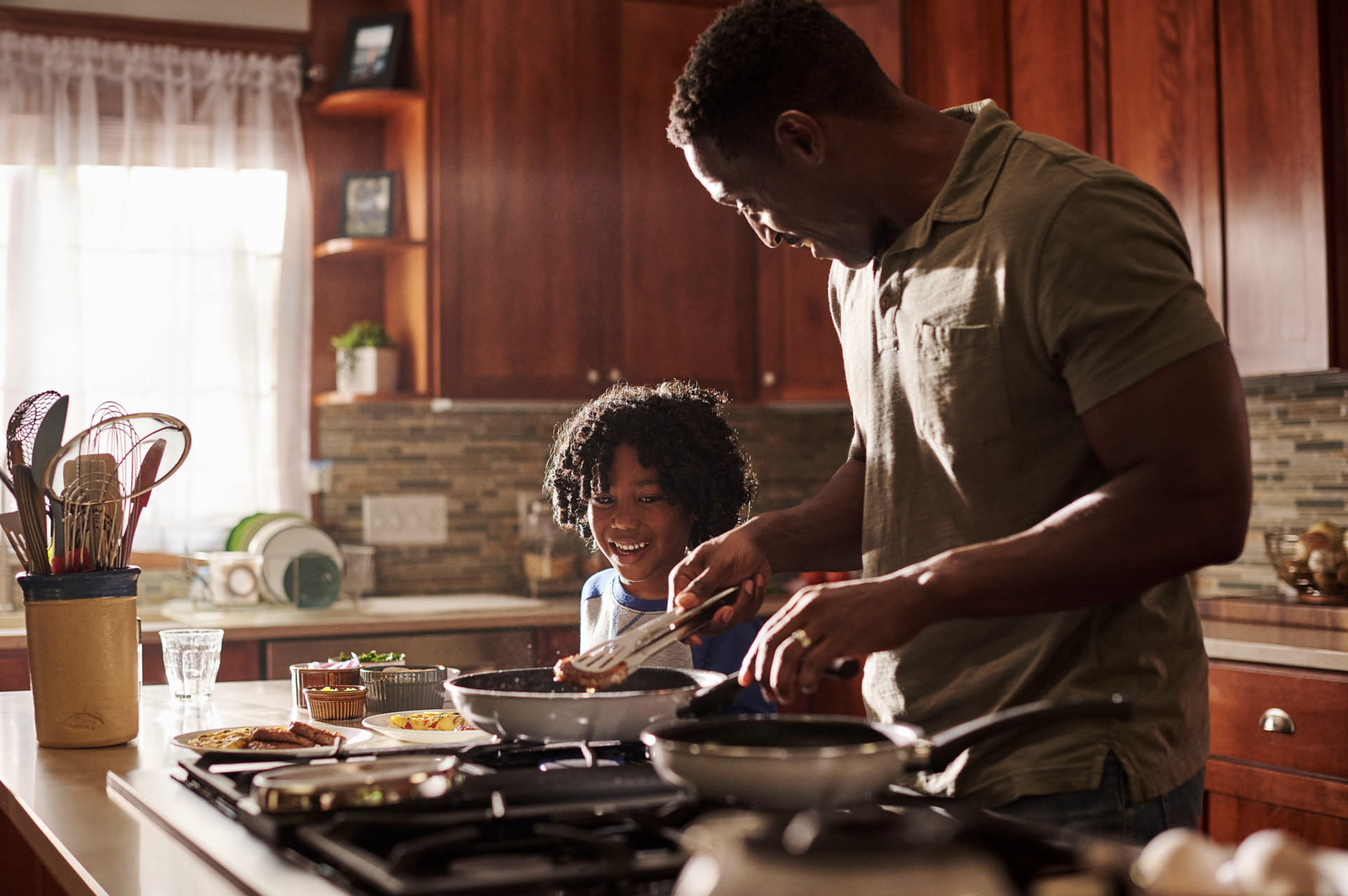 African American father and child cooking together at a kitchen stove, preparing food in pans for a meal at home indoors.