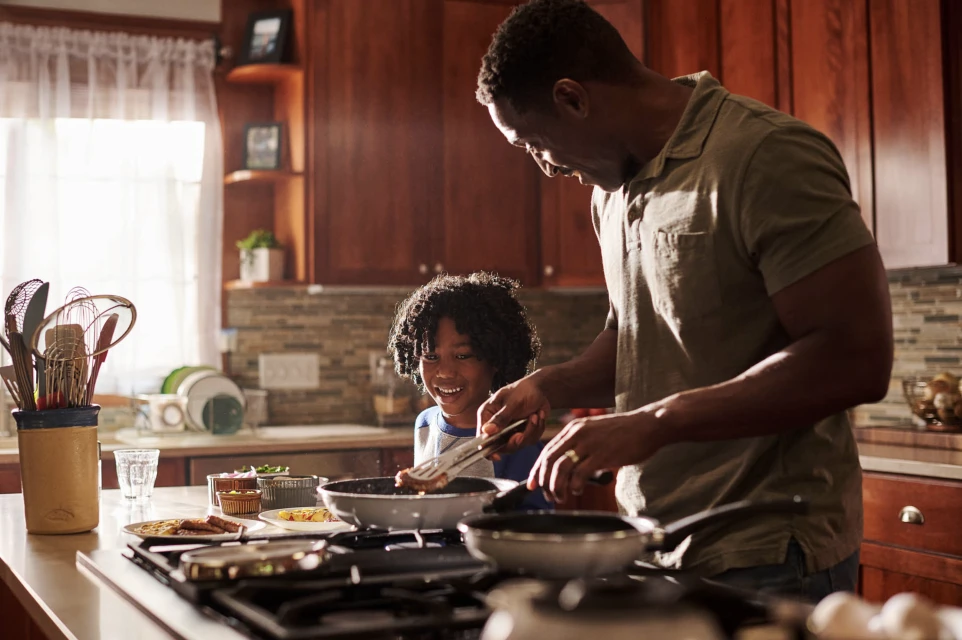 African American father and child cooking together at a kitchen stove, preparing food in pans for a meal at home indoors.