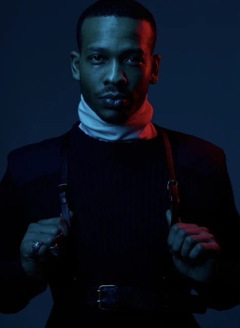 Portrait of a fashionable adult man with a serious expression, wearing a black turtleneck sweater and leather harness, lit with blue and red.