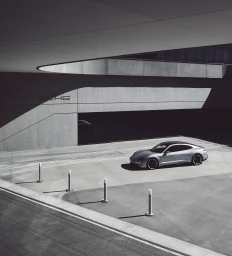 Silver electric luxury car parked in a concrete modern architectural courtyard with harsh sunlight and deep shadows by Anthony Tahlier