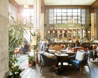 Spacious hotel lounge with patrons seated at tables, a staffed bar in the background, tall windows, and indoor plants.