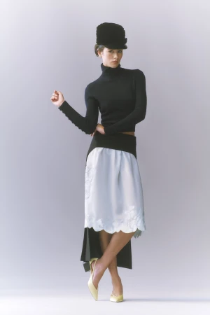 Model in black turtleneck and tall hat wearing a light blue midi skirt against a pale grey studio background