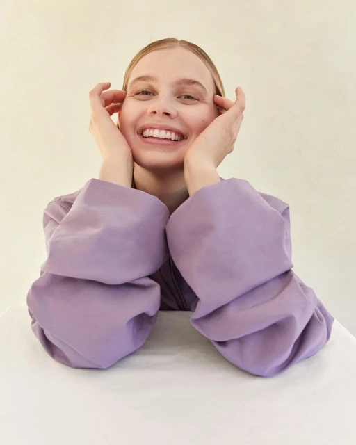 Smiling young woman with hands on her cheeks, wearing a puffy-sleeved lilac top, seated against a neutral beige background. Close-up, portrait.