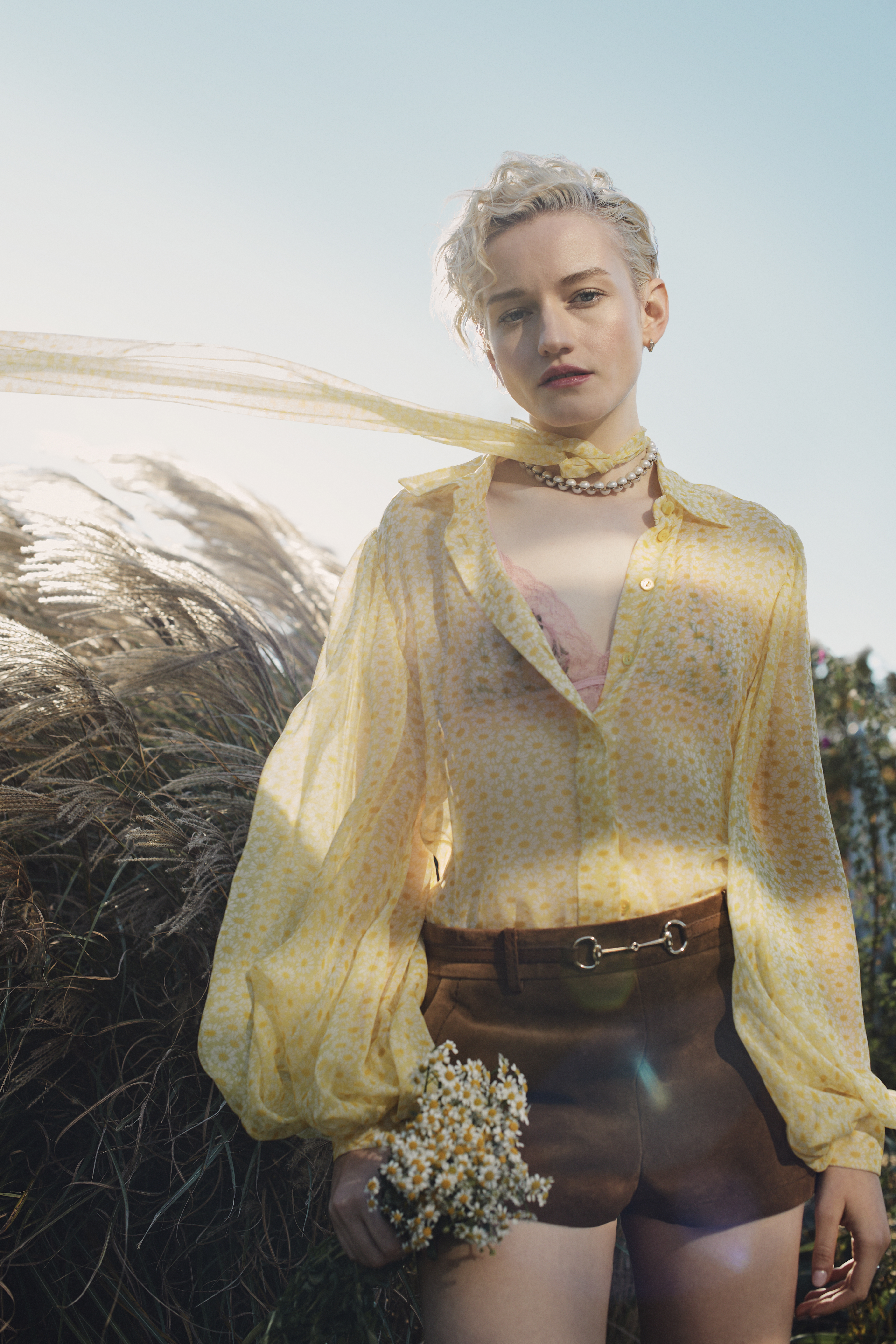 Blonde woman in sheer yellow floral blouse and brown shorts stands in a sunlit grassy field holding daisies, accessorized with pearls.