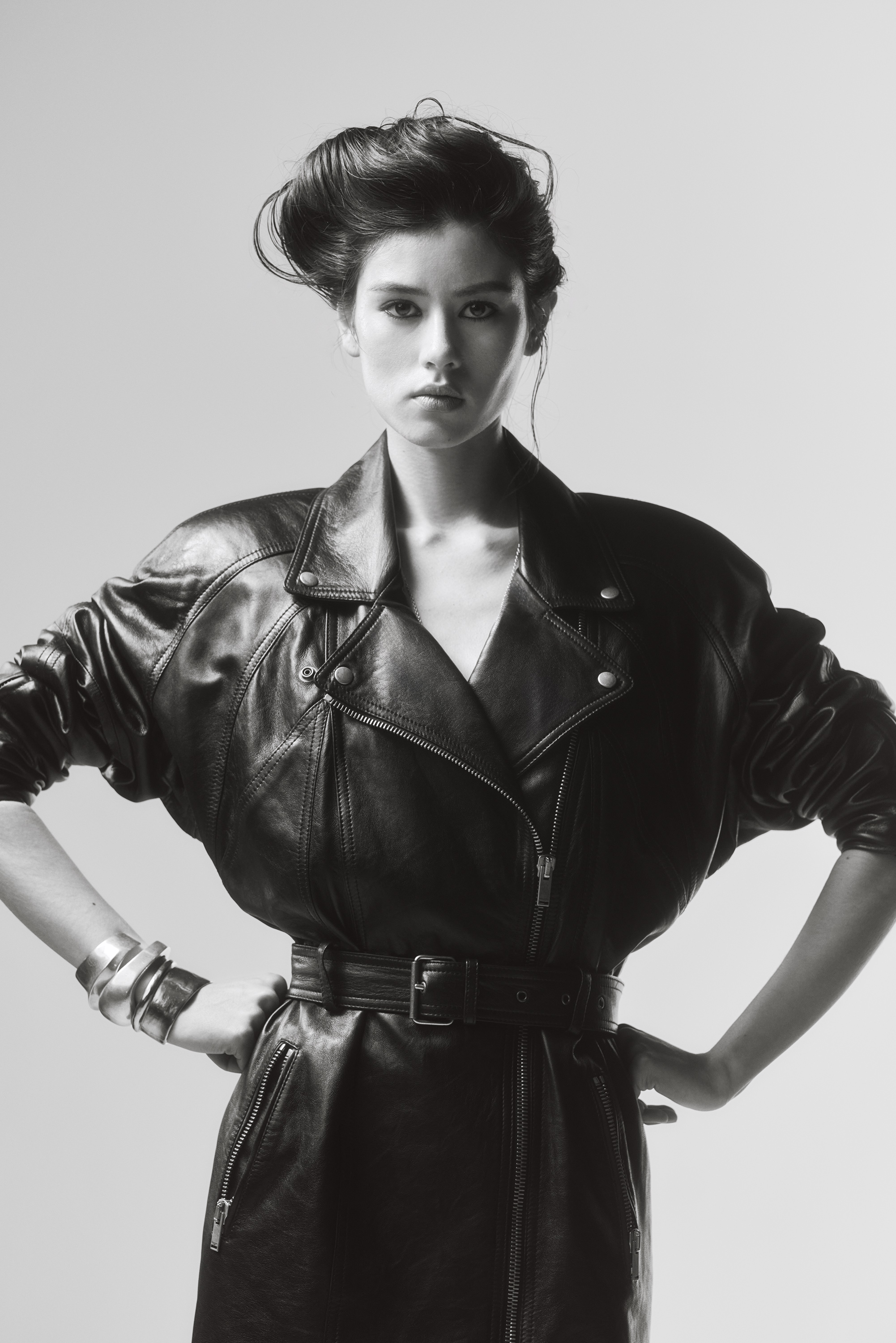 Monochrome portrait of a woman wearing a belted leather jacket with pronounced shoulders against a grey seamless background.