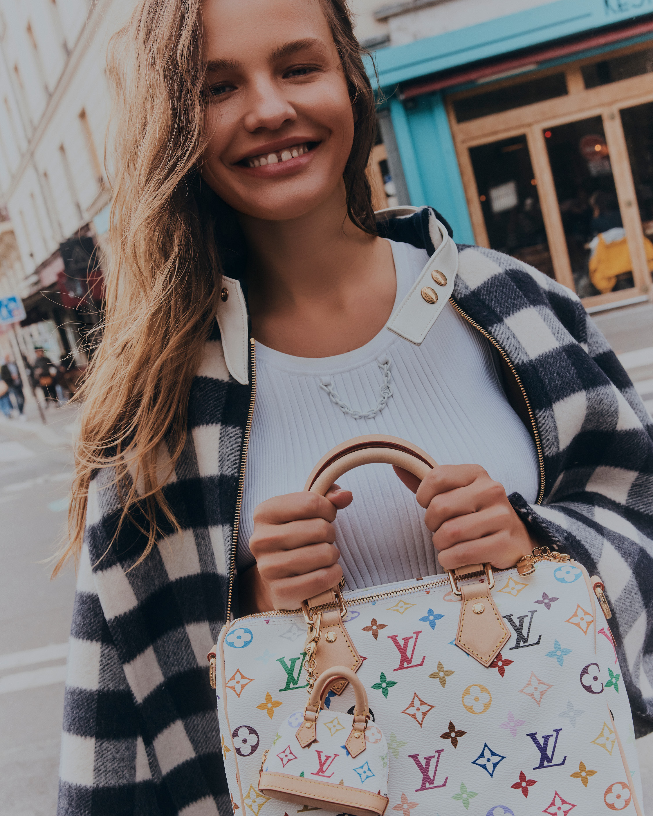 Smiling woman with handbag on city street. Shoulder-length blonde hair, buffalo-check jacket, white top, carrying monogrammed purse.