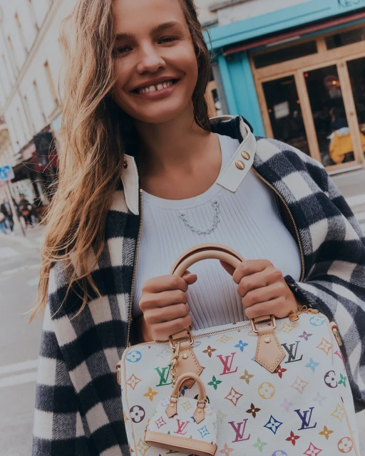 Smiling woman with handbag on city street. Shoulder-length blonde hair, buffalo-check jacket, white top, carrying monogrammed purse.