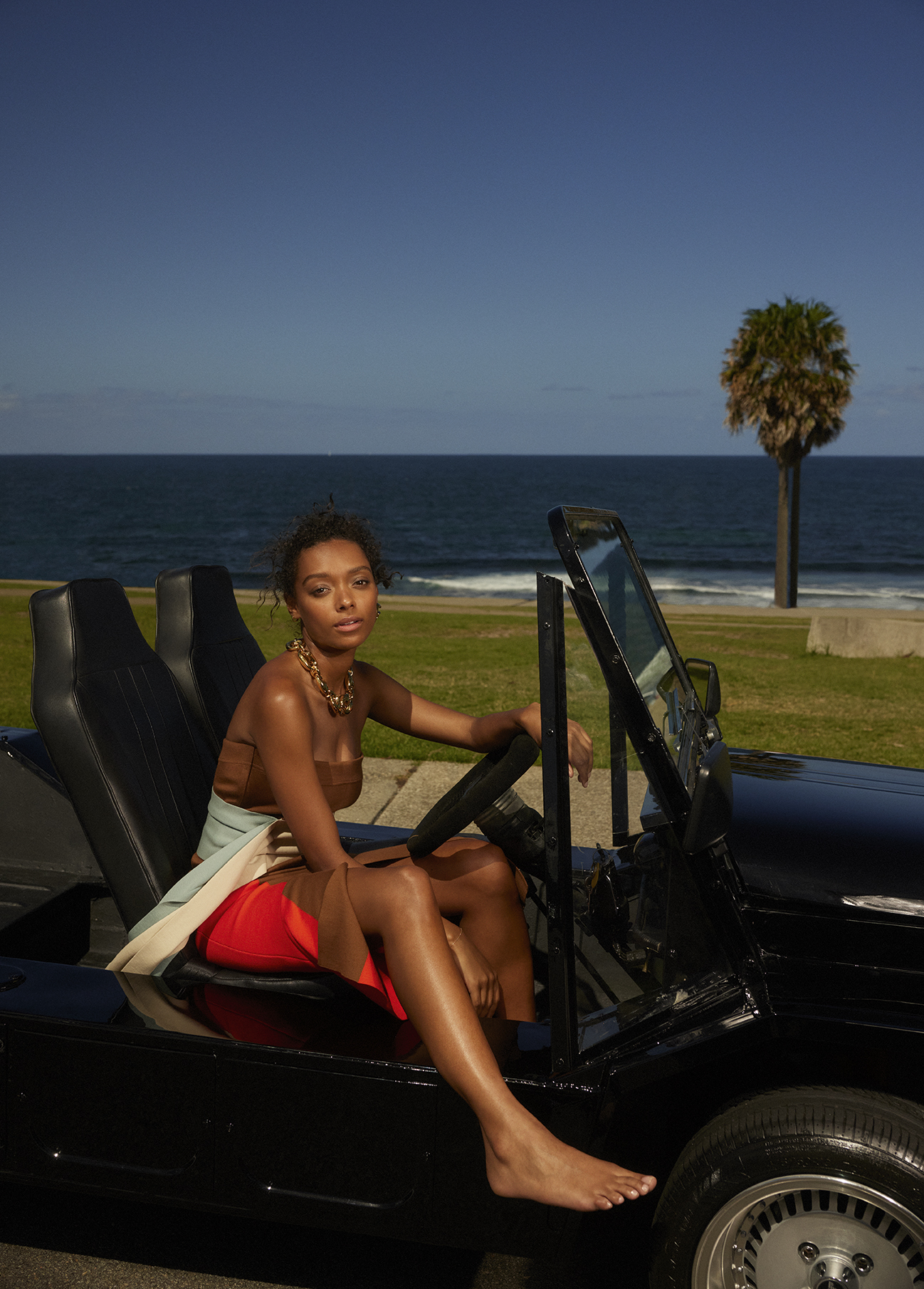 A woman seated in a black convertible car by the ocean, wearing a brown top and red skirt with a gold chain necklace.