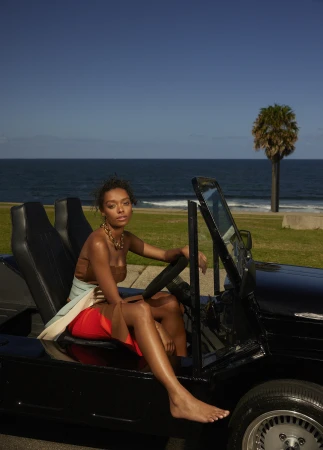 A woman seated in a black convertible car by the ocean, wearing a brown top and red skirt with a gold chain necklace.
