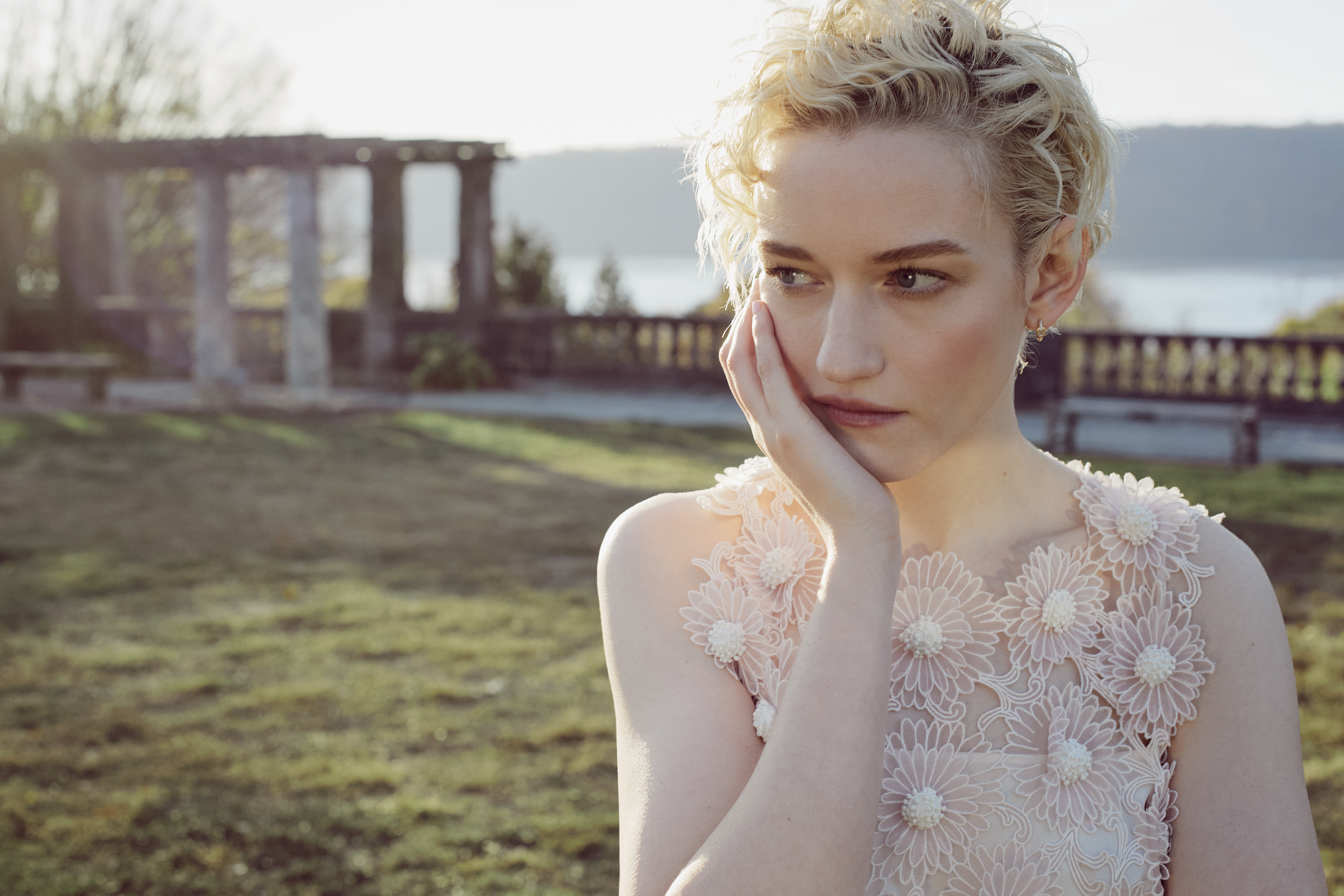 Portrait of a blonde woman in a floral dress outside. She is standing in a grassy field, posing in front of stone structures.