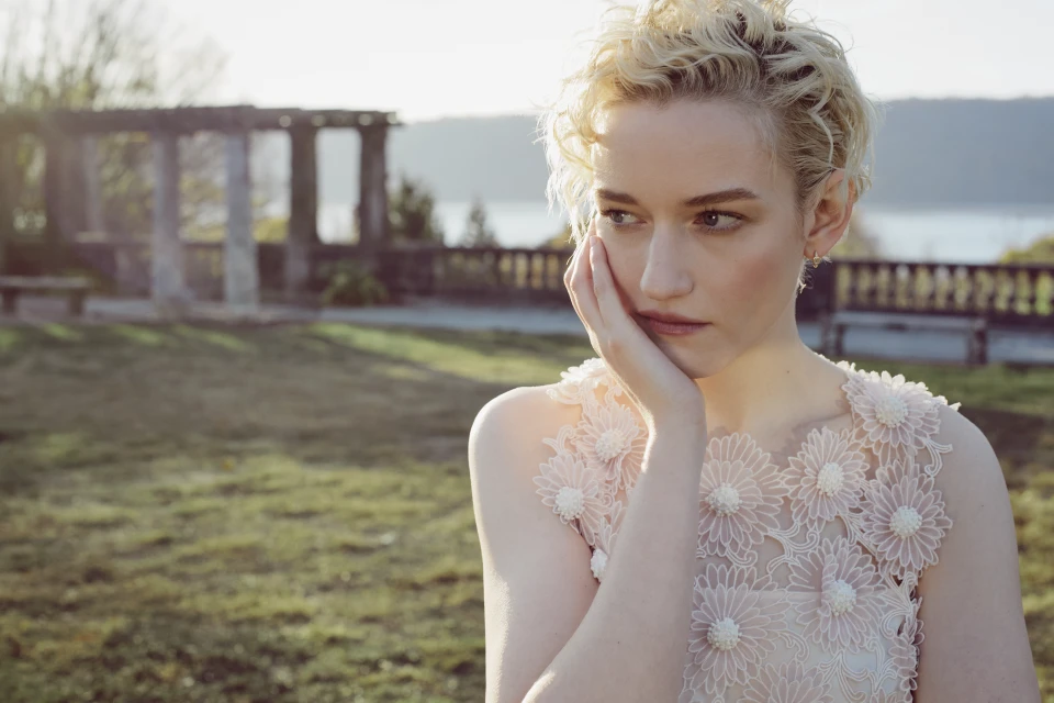 Portrait of a blonde woman in a floral dress outside. She is standing in a grassy field, posing in front of stone structures.