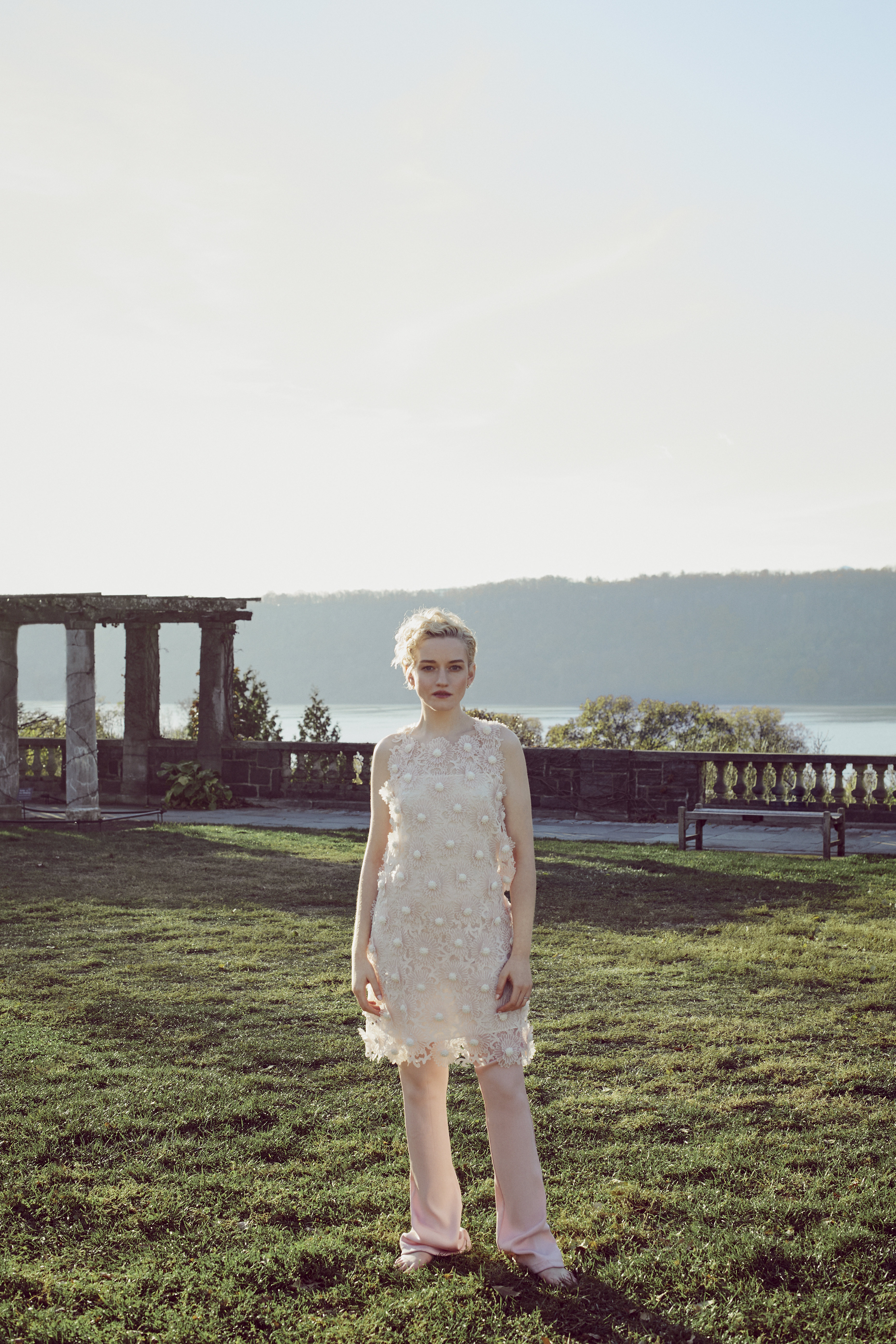 A young woman stands on a grassy lawn wearing a beaded dress and pink pants, with a pergola and lake in the background.