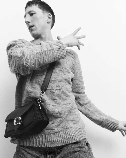 A person in a sweater and jeans poses with a black leather purse, against a white background, in a studio setting. Fashion photography.