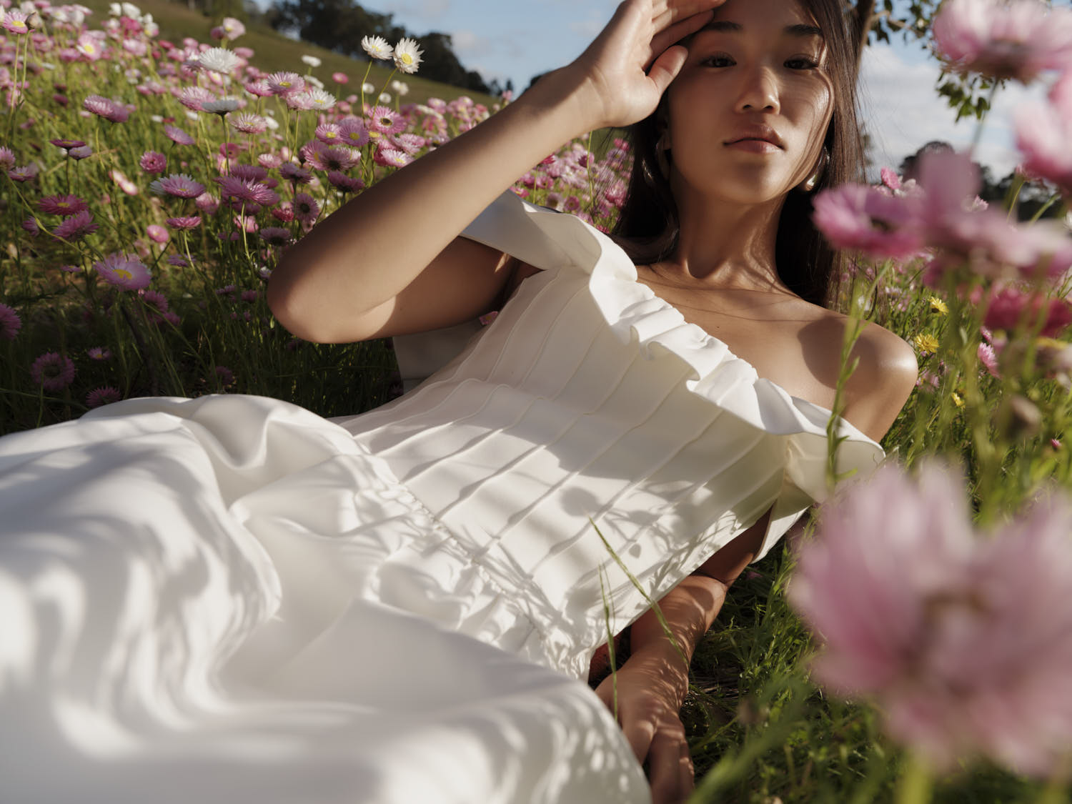 Woman in white dress lays in a field of pink and white daisies with green grass under natural sunlight, eye-level angle. 