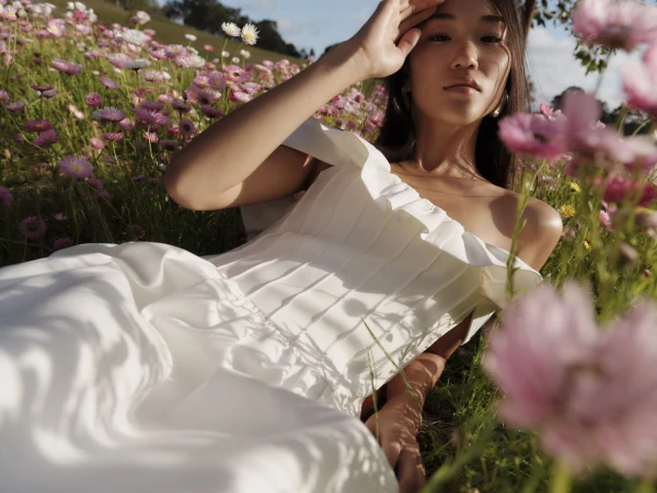 Woman in white dress lays in a field of pink and white daisies with green grass under natural sunlight, eye-level angle. 
