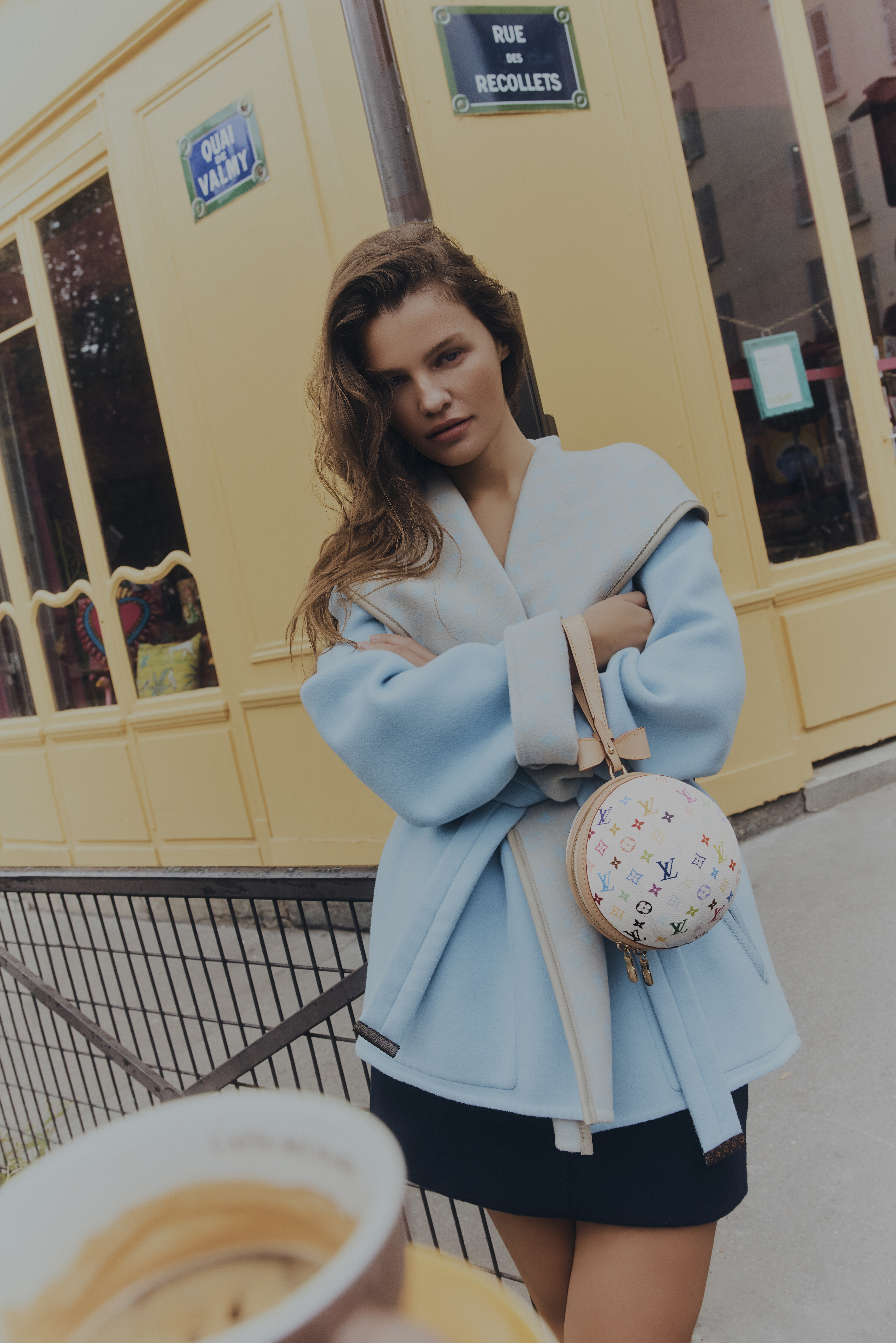 Adult female in an outerwear jacket, posing with a patterned round handbag in an outdoor, urban setting on a daytime shoot.