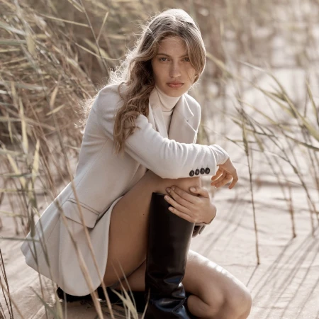 Woman wearing a cream blazer and tall black boots crouching among tall dry grass outdoors.