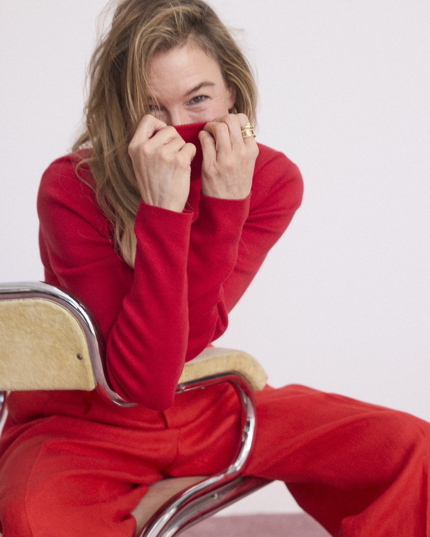 Woman in a red turtleneck sweater pulling the collar up over her face while sitting on a chair, against a white seamless background