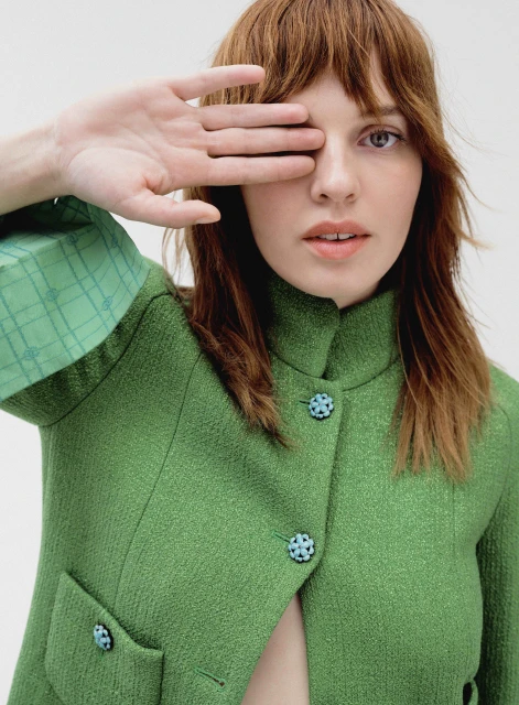 Portrait of a woman with red hair wearing a green tweed jacket, covering one eye with her hand, in a brightly lit studio.