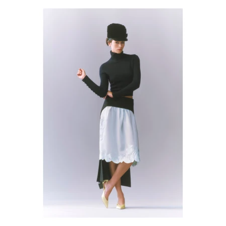 Fashion model in studio poses in a black turtleneck top, pale blue skirt, hat, and yellow shoes, standing against a gray gradient backdrop.