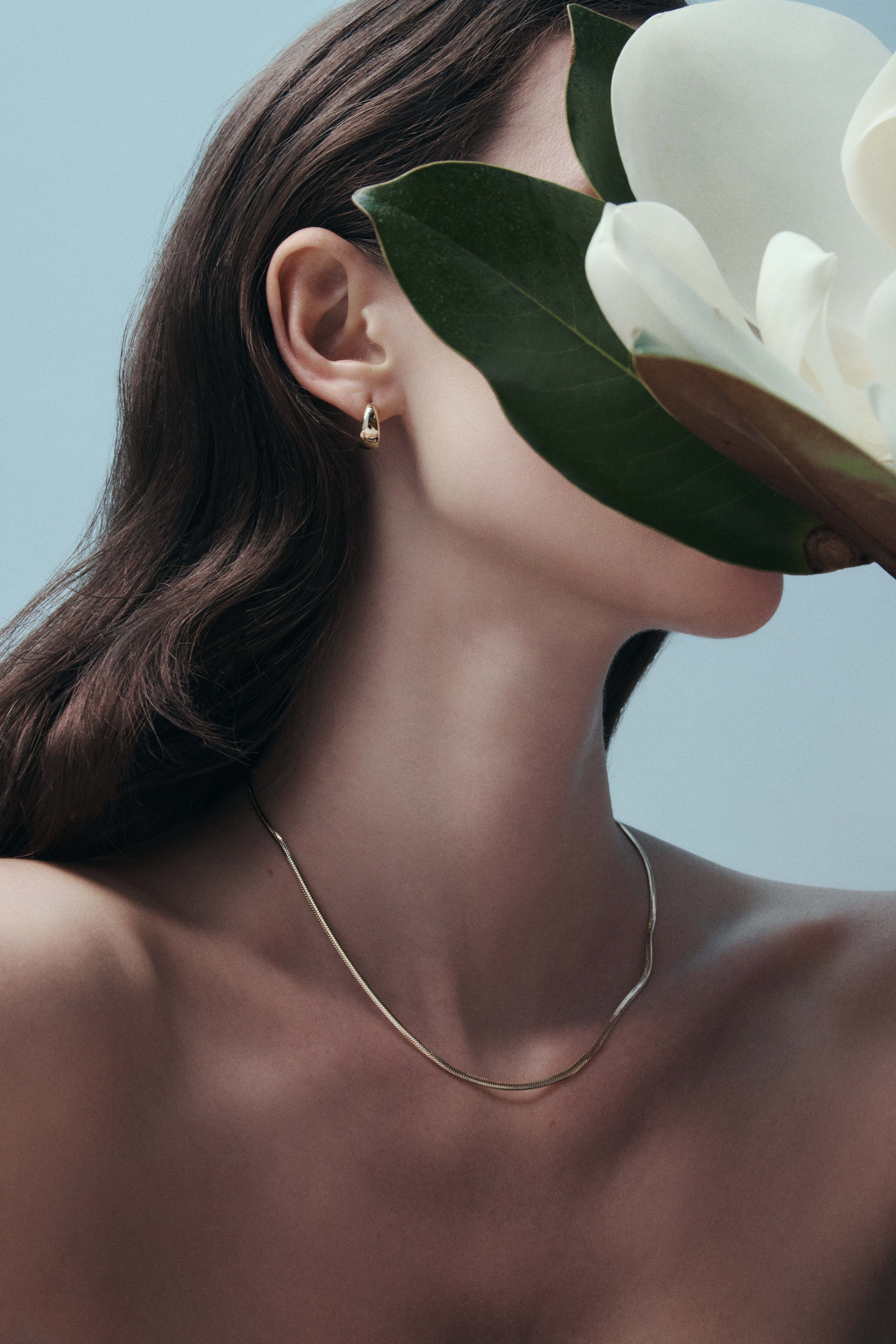 Close-up portrait of a woman holding a large white magnolia flower hiding her face, wearing a gold hoop earring and thin necklace against a pale blue background.