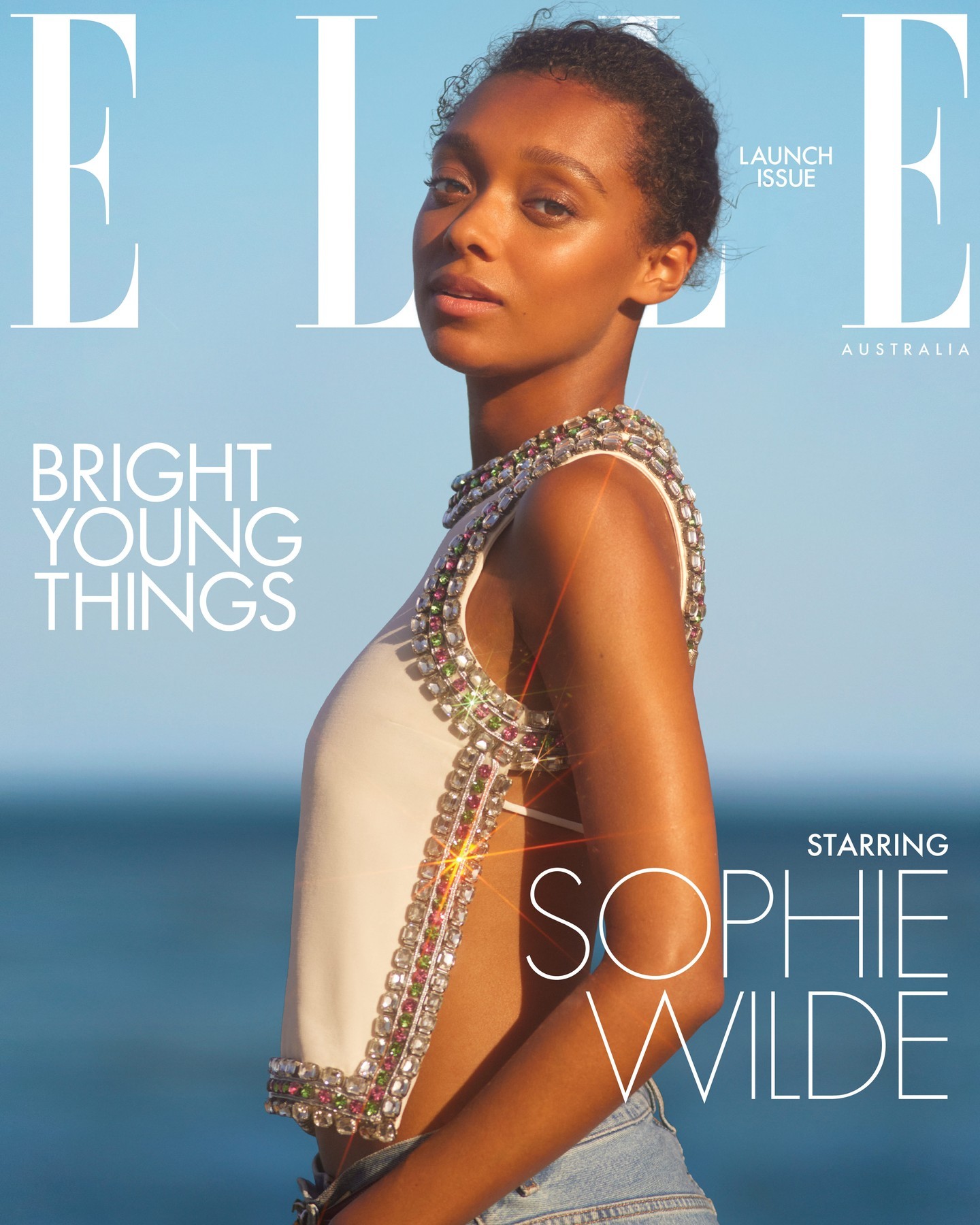 Fashion magazine cover featuring a woman with embellished top posing against an ocean and blue sky backdrop, with 'ELLE' logo.