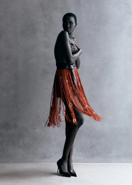 Fashion portrait of an African woman with dark skin, wearing a fringe skirt, posing in a photo studio against a neutral backdrop.