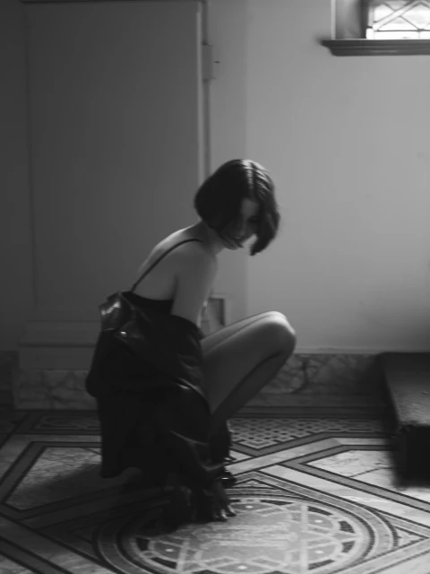 Black and white portrait of a woman in a slip dress and jacket kneeling on a patterned tile floor in front of a closed door.