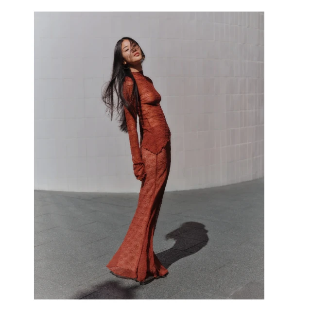 A woman with long dark hair poses in a long-sleeved, reddish-brown, textured dress, standing on a gray surface in front of a light wall.