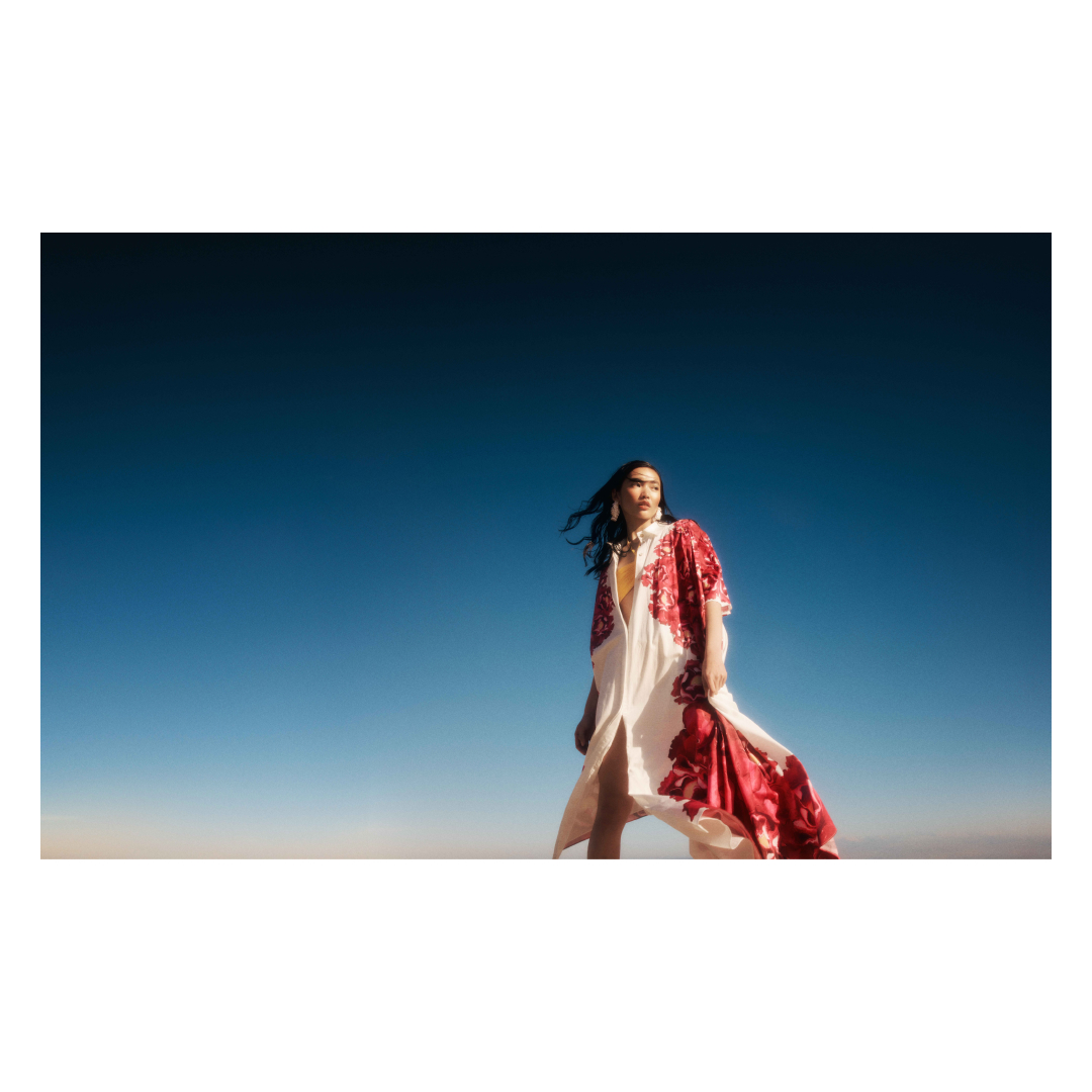 Fashion shot of a woman wearing a white dress with red flower patterns, standing in a desert landscape beneath a bright blue sky.