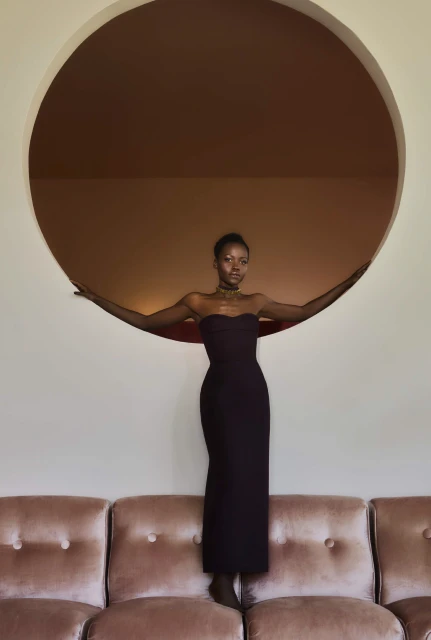 Elegant Black woman in strapless gown poses on vintage couch under round window, creating retro fashion and interior shot.