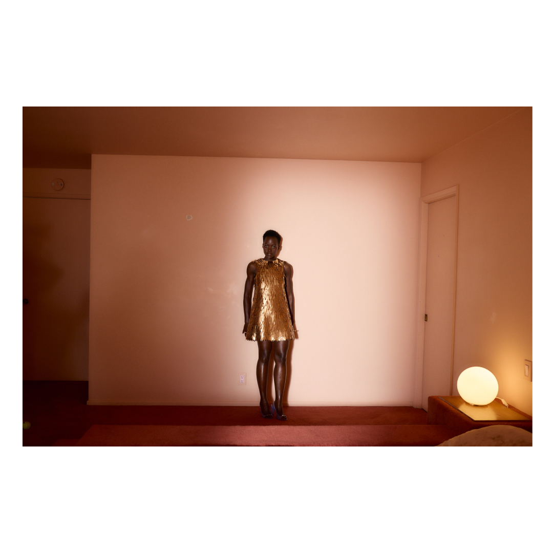 Adult woman in gold sequin dress stands in an indoor space, illuminated by a light on a nearby table, creating a glamorous and elegant look.