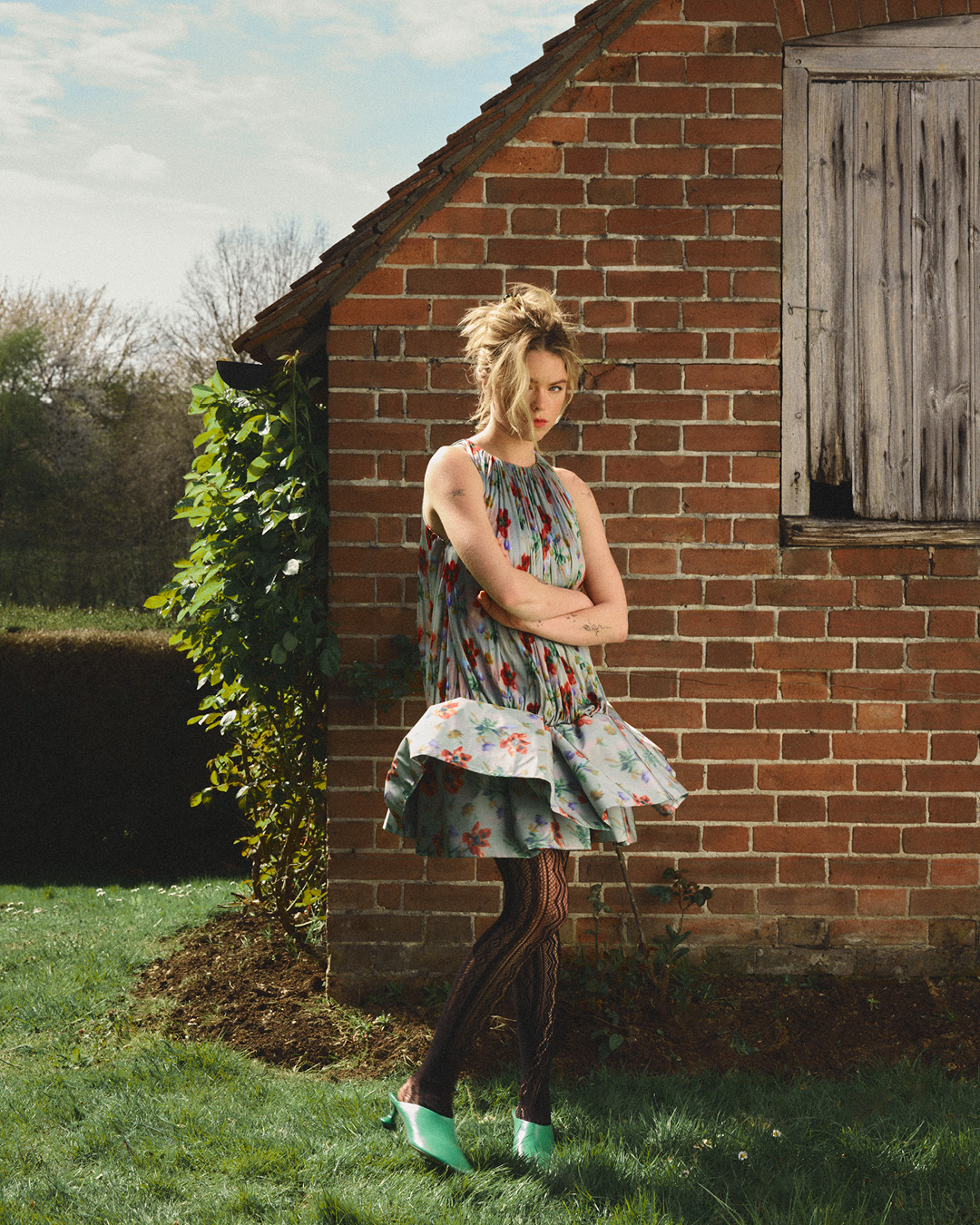 A young woman stands in a floral dress and tights in front of a brick building with greenery, bathed in natural light, in a fashion-editorial shot.