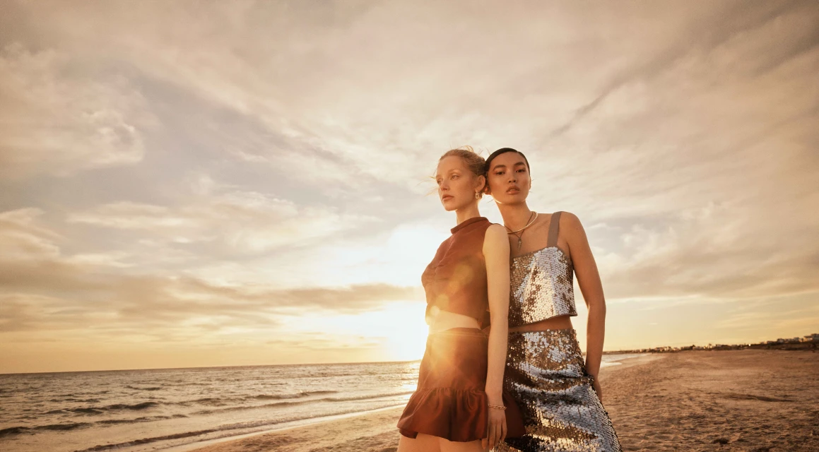 Two women in fashionable attire stand on a beach at sunset, posing against a backdrop of sand, sea, and a sky filled with warm light.