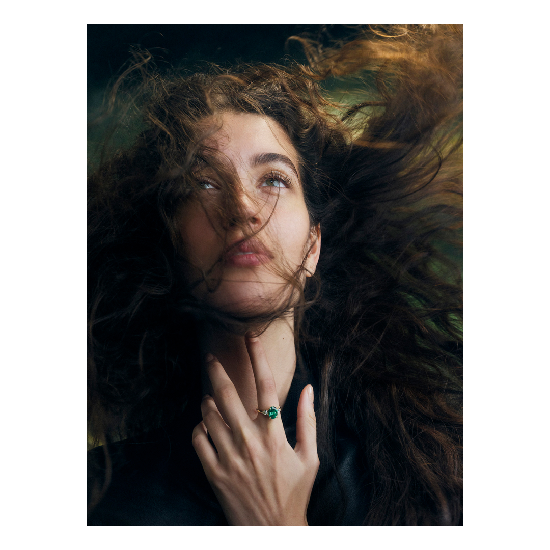 Portrait of a woman with long, dark, curly hair and a gold ring with a green stone on her finger looking upwards in soft lighting.