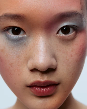 Close-up portrait of a young east asian woman featuring blue eyeshadow and visible freckles.