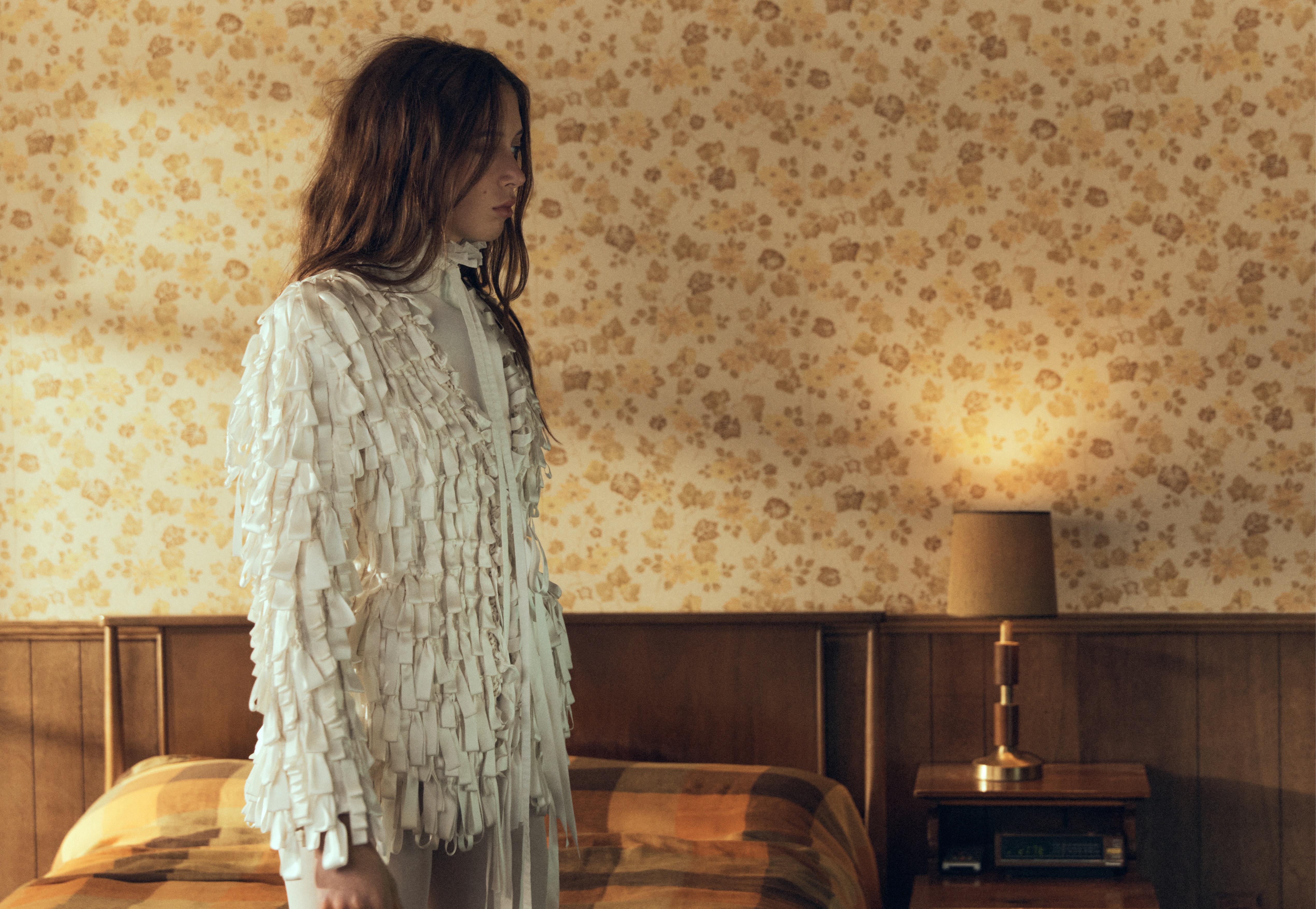 A woman in a white textured fringe jacket stands in profile inside a retro bedroom with floral wallpaper and wooden headboard.