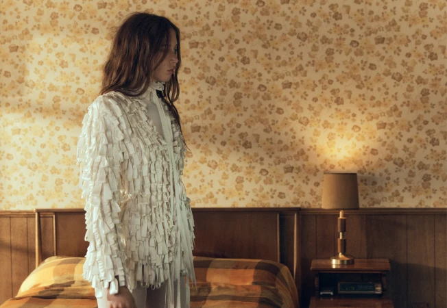 A woman in a white textured fringe jacket stands in profile inside a retro bedroom with floral wallpaper and wooden headboard.