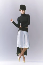 Model in black turtleneck and tall hat wearing a light blue midi skirt against a pale grey studio background