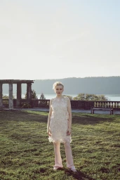 Young blond woman in a cream embellished dress and pink trousers standing on grass before a stone balustrade and river.