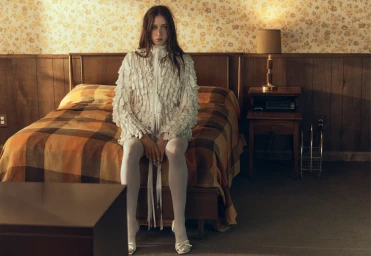 Young woman in white textured ruffled top and tights seated on a plaid bed in a wood-paneled motel room.