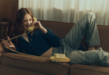 Young woman lying on a brown sofa talking into a retro yellow rotary telephone wearing a navy blue cardigan and jeans.