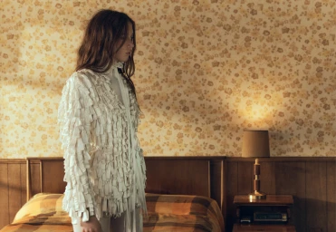 A woman in a cream textured fringe jacket stands in a retro bedroom with floral wallpaper and a wooden bed frame.