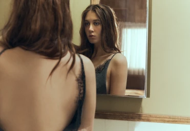 A woman with long brown hair in a black lace slip looking into a bathroom mirror with her back to the camera.
