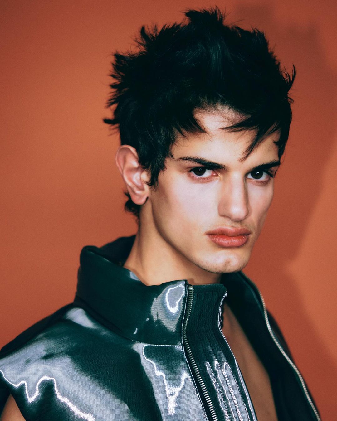A young adult male with short dark hair and a serious expression, wearing a shiny black vest against a solid orange background.